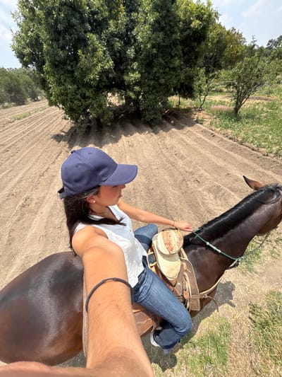 A high-angle selfie depicts a person on horseback in a field near San Andrés Calpan, Mexico. The person wears a navy baseball cap, a white shirt, and blue jeans, and is seated on a dark brown horse with a western saddle. The horse is facing away from the camera. The background features rows of tilled soil and green vegetation. The sky is not visible.