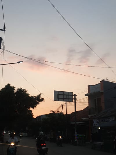 This is a low-angle, vertical photograph taken at dusk in Proyonangg, Indonesia. The sky is a gradient of light grey at the top, transitioning to a soft orange-pink hue near the horizon, suggesting the sun is setting. Wispy clouds are visible, tinged with the colors of the sunset.

The foreground is dominated by a network of dark electrical wires crisscrossing the frame. In the middle ground, silhouetted against the sky, is a street with buildings on the right side. These buildings appear to be shops or residences, with some having multiple stories and visible window frames. One building has a sign with text that is partially visible and difficult to read, but it appears to be in a non-Latin script.

Below the buildings and wires, a street scene unfolds. Several motorbikes are parked or moving along the street. Some have their headlights on, casting a warm glow. One person is visible walking on the street, also illuminated by a nearby light. Another motorbike has a rider wearing a white helmet. Trees with dark foliage frame the right side of the scene, obscuring parts of the buildings and street. A street lamp is visible on the left side of the frame, its light illuminated. The overall impression is a busy street scene transitioning from daylight to evening.