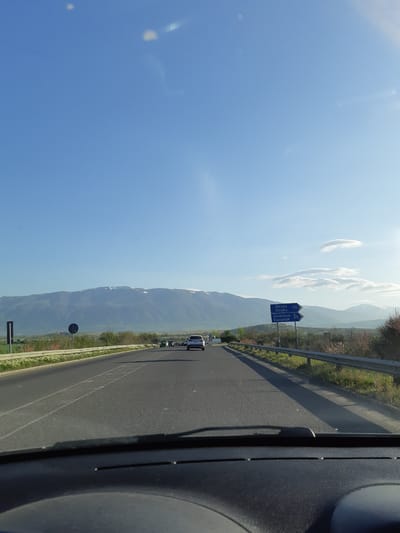 The image captures a bright, sunny daytime view from inside a vehicle traveling on a highway in Bulgaria, consistent with the Marino Pole location. On the right, a dark portion of the car's interior, possibly the dashboard or A-pillar, is visible. The asphalt road stretches into the distance, marked with white lines, with a metal guardrail along its right side.

Beyond the guardrail, green vegetation lines the road. In the mid-ground, a blue road sign provides directions and distances in Bulgarian and English: "Шипка Shipka 70" and "Казанлък Kazanlak 55". Several other vehicles are visible on the road ahead.

The background features a grand mountain range with some snow-dusted peaks, extending across the horizon under a clear, vibrant blue sky dotted with a few elongated white clouds. The scene conveys a sense of a pleasant road trip through a scenic, mountainous landscape under excellent weather conditions.