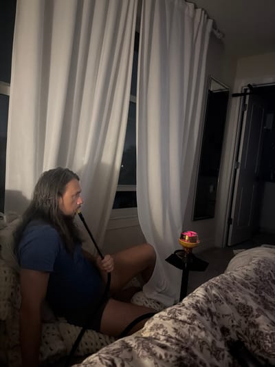 A person with long, wavy hair is sitting on a couch, holding a hookah hose and smoking. The person is wearing a blue t-shirt and dark shorts. The room is dimly lit, with white curtains covering a window. A small black table with a lit hookah is beside the couch, and a bed with a floral patterned comforter is visible in the background. The scene appears to be an indoor residential setting in Lauderdale Lakes, Florida.
