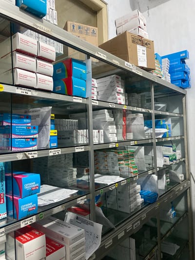 The image shows a pharmacy shelf stocked with various medications. The shelves are made of metal and glass, and they are organized with labels indicating the names of the medications. On the shelves, there are boxes of Atorvastatin Calcium Trihydrate, Amoxicillin Trihydrate, Allopurinol, Cefixime Trihydrate, and Cefadroxil Monohydrate. There are also some larger boxes and stacks of smaller boxes. The overall impression is one of a well-stocked pharmacy. The presence of labels in Indonesian, such as "Kapsul," "Tablet salut selaput," and the name of the pharmacy "RUMAH SAKIT ISLAM PATI," indicate that this is a pharmacy in Indonesia. A sign on one of the shelves reads "RG-09", which could be an inventory code. The image is a medium shot, taken from a slightly low angle, showing the shelves filled with medicines.
