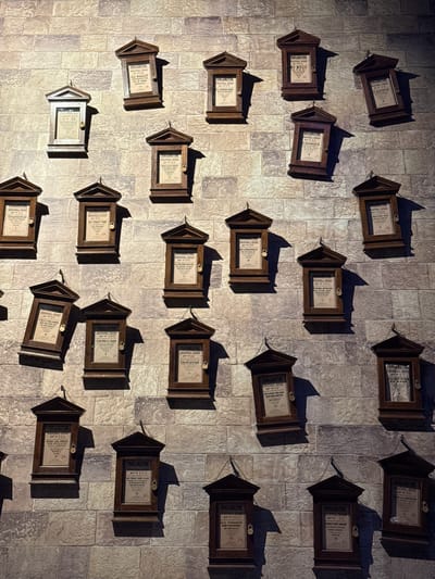 The image shows a wall covered in stone-like tiles. Attached to the wall are numerous wooden frames with small doors, similar to small cabinets or lockers. Each frame contains a printed document, likely proclamations or decrees. The documents have visible text, though it's largely obscured, some words like "PROCLAMATION," "EDUCATIONAL DECREE," "ACTIVITIES," and "STUDENT" are discernible. The frames are scattered randomly across the wall, casting shadows. The scene suggests a specific setting, possibly a themed area in a museum or an attraction based on a fantasy world, where rules or regulations are posted in an old-fashioned manner. The city of Nerima, Japan, is not explicitly discernible.