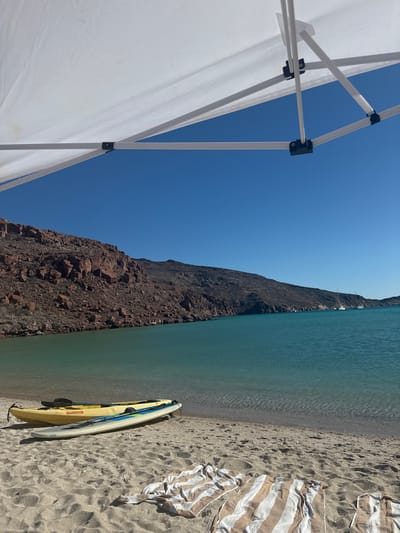 On a sunny day in Unknown, Mexico, a beach scene unfolds. The azure sea meets a sandy shore lined with a rugged, dark-colored cliff face. A yellow and white kayak rests on the sand near the water's edge, adjacent to a striped towel. In the foreground, the corner of a white canopy tent is visible. The sky is clear and blue, and several boats are visible in the distance.