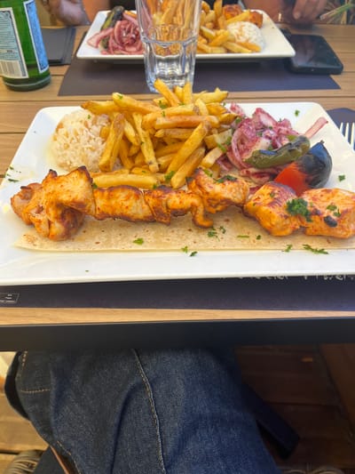 This detailed image depicts an outdoor dining scene in Saint-Just-Saint-Rambert, France. The primary focus is a white rectangular plate in the foreground, presenting a meal consisting of a chicken shish kebab, a mound of white rice, a generous portion of golden french fries, a side salad of thinly sliced red onions, and two pieces of grilled vegetables (one green pepper, one red pepper) with charred skins. The chicken pieces on the skewer are golden-brown with visible grill marks and are garnished with chopped green herbs, resting on a flatbread. The plate is situated on a dark placemat, which displays a "MIX Paper FSC® C002321" label, over a slatted wooden table. Below the table, a person's legs clad in dark blue denim jeans are visible.

In the midground, a green glass bottle is positioned to the upper left. A second white rectangular plate is partially visible, containing french fries, rice, and red onion salad, suggesting another similar meal. Two individuals are partially visible in the background: one's hand near the green bottle on the left, and another's hand on the right holding a dark-colored smartphone over the table. The tables are constructed from light-colored wooden slats, and the scene is brightly lit, consistent with daytime.