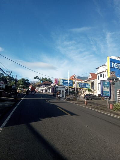 The image captures a wide-angle view of a two-lane asphalt road in Khouathineung, Laos, under a clear blue sky sparsely dotted with thin white clouds. Bright sunlight illuminates the scene, casting elongated shadows from unseen overhead objects onto the road surface, suggesting either early morning or late afternoon.
The road features a solid white line delineating its left edge and a segment of a double solid yellow line marking the center further into the distance. On the left side, a sloped embankment and retaining wall support dense green foliage, several utility poles, and an intricate network of overhead electrical wires. A red circular sign marked "OUT" is partially visible among the trees. Multiple motorcycles, some stationary, are present in the left lane in the mid-ground, with several individuals visible near them. A brown truck is partially observable further down this lane.
The right side of the road is bordered by a series of commercial buildings and structures. A dark-colored SUV is parked on the roadside, adjacent to a stone-patterned wall. Above this, a large blue and yellow sign prominently displays "DELAGA BIRU Convention & Hote," with a smaller sign below indicating "START FROM RP 235K." A white booth-like structure stands further along the right, preceding additional buildings with red-tiled roofs. An orange traffic cone is positioned on the roadside near the parked SUV. A single individual, wearing a dark jacket and helmet, operates a white scooter in the right lane, moving away from the viewer.
Distant mountains or large hills are visible on the horizon, partially obscured by the foreground structures and vegetation.