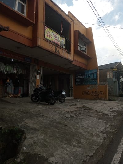 The scene depicts a street view in Ura-Dozhi, Bhutan, featuring a two-story commercial-residential building under a partly cloudy sky. The building, predominantly orange-yellow with darker brown trim, shows signs of weathering and staining. Its ground floor features an open storefront on the left, displaying various packaged goods, with two individuals partially visible inside; one in a light garment, facing away, and another in a dark shirt. A partial sign reading "BUKA SETIAP HARI" is visible above this shop. To its right are two closed metal roll-down gates. The upper floor includes a closed window on the left, an open balcony with a black metal railing displaying a commercial banner (featuring text and images) and a red and white striped flag, and an open window alcove to the right. A blue sign featuring an image resembling ancient ruins and text including "GARDEN CIPADU" is affixed to the lower section of the building's right side, below which "GRAB ON ROAD -Fahmi Dava" is graffitied in black. Two dark-colored motorcycles are parked on the uneven, grey concrete foreground in front of the closed gates. The concrete surface exhibits cracks and discoloration, with a small patch of greenery visible in the bottom-left. Utility lines crisscross the sky. In the background, to the right, a single-story brick building with a dark roof is visible.