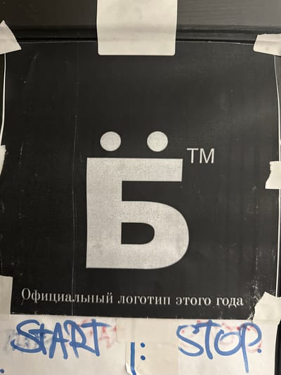 The image shows a black sign with a stylized silver-colored logo resembling a "5" with two dots to the left, and the letters "TM" (trademark symbol) above it. To the right of the logo is text in what appears to be Russian, vertically aligned. The sign is partially covered with pieces of white tape on which the words "START" and "STOP" are written in blue marker. The sign appears to be affixed to a dark-colored surface with a black border. The image has been rotated to show the sign on its side. The city is Новоглаголево, Russia.