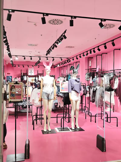 The image depicts the interior of a brightly lit clothing store with predominantly pink walls and flooring. Two mannequins are prominently displayed at the front, both wearing novelty animal-ear masks and fashionable outfits. The mannequin on the left is wearing a white lace top and denim shorts, while the mannequin on the right is in a purple t-shirt and white skirt. Behind the mannequins, clothing racks filled with various garments are visible, illuminated by numerous black track lights mounted on the ceiling. A promotional poster advertising a "50% ONE PRICE" sale is displayed near the left mannequin. The store appears to be located in the city of Nola, Italy, as indicated by text on a sign that reads "NUOVA COLLABORAZIONE" and mentions "NOLA".