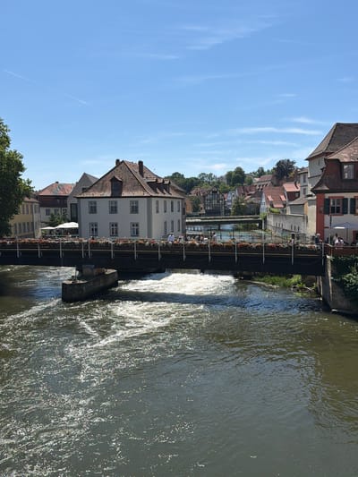 The image depicts a sunny day in Bamberg, Germany, likely along the Regnitz River. In the foreground, a body of water flows energetically, with sunlight glinting off its surface and creating white frothy patterns. A concrete structure, possibly part of a weir or bridge support, is partially submerged in the water.
Spanning across the river is a bridge adorned with numerous planters overflowing with colorful flowers. People are visible on the bridge, some walking and others appearing to be seated at an outdoor cafe to the right. The bridge connects to a street lined with traditional German buildings.
The architecture features pitched red-tiled roofs, white facades, and multi-paned windows. Some buildings have timber-framed elements, characteristic of historic German towns. In the background, more buildings ascend a gentle slope, interspersed with trees, and a second, smaller bridge is visible further upstream. The sky is a clear, bright blue with a faint contrail from an airplane stretching across it. The overall scene conveys a vibrant and picturesque urban landscape.