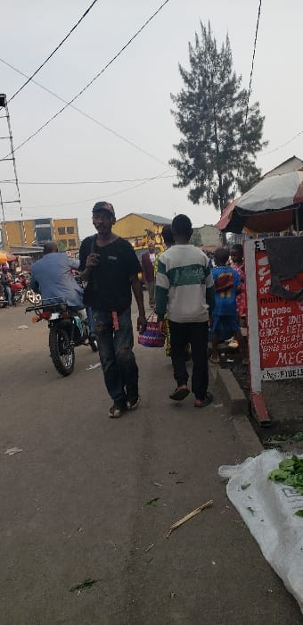An outdoor street scene in Buzove, Democratic Republic of the Congo, captured under an overcast sky. In the foreground, a male individual wearing a dark t-shirt, ripped blue jeans, and a baseball cap walks towards the viewer, with a dark strap across his left shoulder. To his right, another male, dressed in a white, green, and black striped long-sleeve shirt and dark pants, walks away from the viewer, carrying a multi-colored woven basket.

To the left of the frame, a male is seated on a dark-colored motorcycle, facing away from the viewer. On the right, a raised concrete sidewalk features a market stall with a red and white striped umbrella canopy. A white sign affixed to a structure displays partial text including "M-pass," "VENTE UNITAIRES," and "MEG." Several individuals, including children, are visible on the sidewalk near the market stall. A large white sack, containing green leafy produce, is positioned on the ground adjacent to the sidewalk.

The ground surface consists of a mix of dirt and pavement, with scattered debris such as leaves and small sticks. In the midground and background, various multi-story buildings are visible. A tall, slender conifer tree stands prominently against the sky. Numerous overhead power lines traverse the upper portion of the frame. The scene indicates a busy urban commercial or residential area with ongoing daily activities.