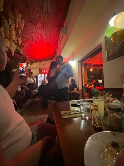 The image shows an interior scene, possibly a restaurant or bar, with patrons. A couple is dancing or embracing in the center. Other people are seated at tables, some using their phones. The room has a red-lit wall on one side with floral decorations. A table with drinks and dishes is in the foreground. Balloons are visible near the ceiling. The location is inferred to be Panagyurski kolonii, Bulgaria.