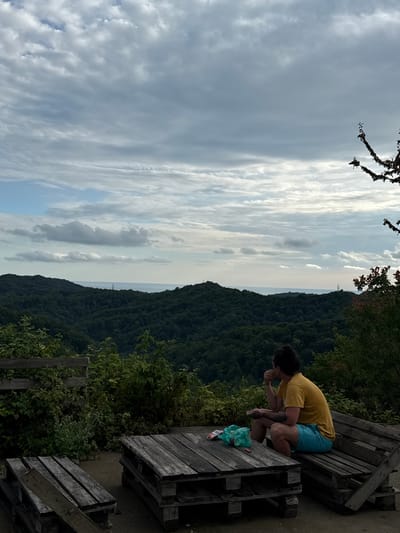 A person is crouching on a wooden bench, leaning forward with their hands near their mouth. They are wearing a yellow t-shirt and blue shorts. The bench is made of weathered wood and appears to be part of a picnic table. To the right of the bench, a green plastic bag is visible.

The setting is outdoors, overlooking a densely forested mountainside that slopes down towards a body of water, possibly the sea, in the distance. The sky is filled with dramatic clouds, suggesting either an overcast day or a time near sunrise or sunset. The overall mood appears contemplative or relaxed. The image is oriented vertically, with the sky taking up a significant portion of the left side. A distant tower can be seen on the horizon.