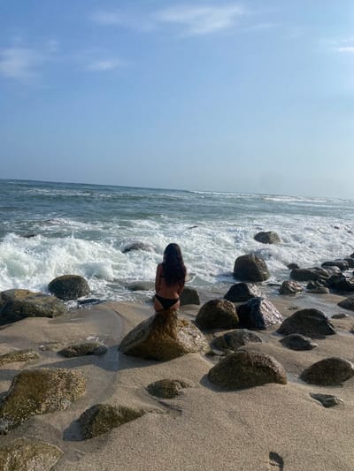 A young woman is sitting on a large rock on a sandy beach in Punta Hermosa, Peru, at coordinates -12.325 latitude and -76.835 longitude. She is wearing a black bikini and has her back to the camera. She is facing the ocean, which is in the background. The ocean is blue and choppy, with whitecaps. The sky is blue and clear. There are several other large rocks on the beach. The sand is light brown and there is a slight shadow cast by the woman onto the rock she is sitting on. The scene is peaceful and tranquil.