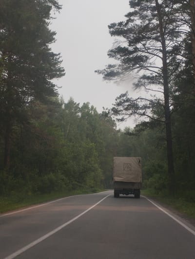 An overcast, foggy day in a forest area near Sosnovka, Russia. The scene is dominated by tall pine trees lining a paved road. A single lane road curves gently to the right and is marked with a white line. In the distance, a canvas-covered truck is traveling away from the viewer down the road. The truck has a logo on its side with Cyrillic lettering. The surrounding environment is dense with trees and foliage, giving a sense of isolation. The light is diffused due to the fog.