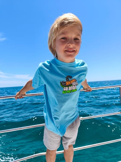 A young boy with blonde hair and fair skin smiles at the camera while standing on a boat. He is wearing a light blue t-shirt with a graphic of a monster truck and the text "HOT WHEELS MONSTER TRUCKS GO BIG! GO HOT WHEELS!". He is also wearing grey shorts. The boy is holding onto a metal railing on the side of the boat. The background shows the bright blue ocean under a clear blue sky. Sunlight is glinting off the water, suggesting it is a sunny day, likely mid-morning or afternoon. The gentle waves indicate a calm sea. The location context is Albufeira, Portugal. The overall mood is cheerful and adventurous.