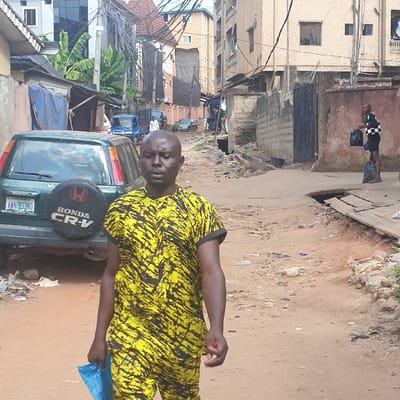 This image captures a daytime scene on an unpaved urban street in Awada, Nigeria. In the foreground, slightly left of center, a bald adult male with a medium-dark complexion walks towards the viewer's left. He wears a short-sleeved yellow and black patterned two-piece outfit and holds a crumpled light blue plastic item in his left hand.

To his immediate left, a dark green Honda CR-V (license plate AWK-933ND) is parked facing away from the camera, its rear-mounted spare tire visible. Along the left side of the street, low structures with corrugated metal or tarp roofs are visible, interspersed with green foliage. Overhead, a dense network of electrical or communication cables spans between utility poles and across buildings.

Further down the street on the right, a slender individual in a black and white checkered shirt and dark shorts stands near a concrete wall, holding two black plastic bags. Adjacent to this individual, an open drainage ditch runs along the right side of the street, partially covered by wooden planks. The road surface is reddish-brown dirt, uneven, and littered with various debris including plastic bags and wrappers.

In the background, multi-story concrete buildings with windows and balconies line the street, indicating a residential or mixed-use area. A light blue three-wheeled vehicle and other cars are visible further in the distance.