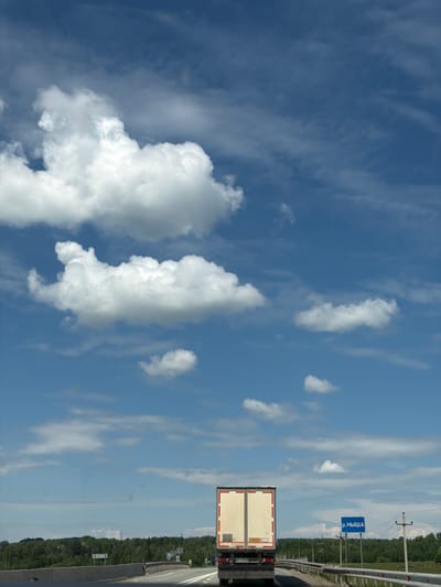An overhead shot captures a semi-trailer truck driving on a highway under a blue sky filled with white clouds. The truck is white with red accents and is positioned in the lower center of the frame. To the right of the truck, a blue sign with white lettering reads "р.НЫША", which translates to "River Nysha". The highway has a concrete barrier on the right side and is bordered by a line of trees. The overall scene depicts a sunny day on a rural highway in Russia, possibly near a river.