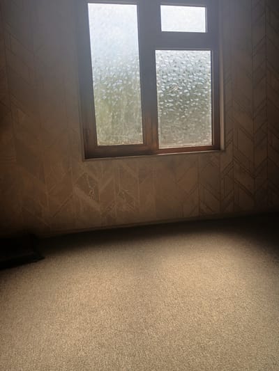 The image shows an interior room with patterned wallpaper and a carpeted floor. A wooden window with textured glass is visible on the back wall. The lighting suggests it is daytime, with sunlight streaming through the window and creating a bright spot on the floor. No people or specific actions are depicted. The scene appears to be a typical domestic interior. The location of Kushkayragach, Uzbekistan, is not discernible from the visual information presented in the image.