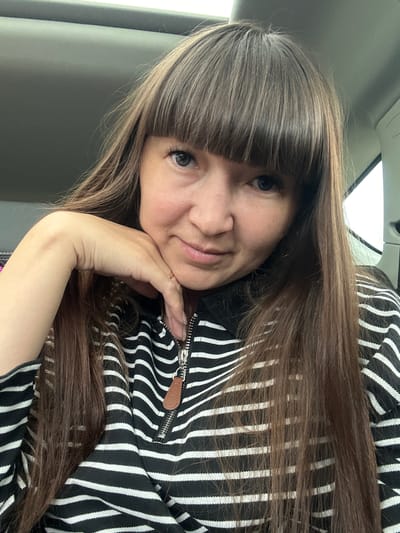 A woman with long brown hair and bangs is looking at the camera, resting her chin on her hand. She is wearing a black and white striped sweatshirt with a zipper. The image appears to be taken inside a vehicle, indicated by the car's interior visible in the background. There are no visible landmarks that would place the location specifically in Dokshitsa, Russia.