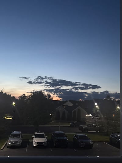 This is a full-frame, eye-level, outdoor, vertical photograph of a residential area at dusk in Allen Township, United States of America. The sky is a deep blue, fading to lighter shades of blue and orange at the horizon, with dark clouds scattered across. Silhouetted against the sky are trees and the roofs of houses. In the foreground, several cars are parked in a lot. From left to right, these include a silver SUV, a white sedan, a black sedan, and a black truck. There are also a few other cars visible further back, including a white SUV driving on a road. Streetlights are on, casting illumination on the parking lot and surrounding area. The scene appears peaceful and quiet, with no people visible.