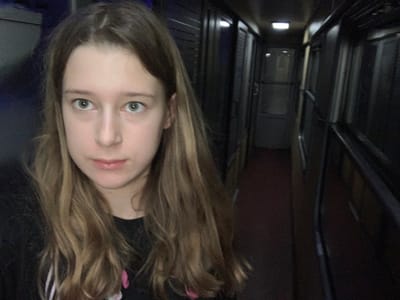 An upside-down portrait of a young woman with shoulder-length blonde hair and blue eyes. The background suggests an enclosed hallway with metallic walls and a door at the far end. The woman's face is central, with her hair framing the top of the image. There are hair accessories on the top part of her head. The lighting in the hallway is dim, with bright spots near the floor. The overall impression is surreal and distorted due to the inverted perspective.