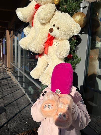 An individual is depicted outdoors in Mistolovo, Russia, positioned in front of a building facade. The subject is partially visible from the chest upward, oriented with their back towards a glass storefront. They wear a bright pink ribbed knit beanie featuring a white circular Moncler logo patch on the front. A light pink puffer jacket covers their torso. The subject's hands are encased in large, fluffy, peach-colored mittens, which are adorned with cartoonish smiling faces, small bunny-like ears, and metallic-toned eyes.
Behind the individual, three large, cream-colored plush teddy bears are suspended vertically, forming a decorative arrangement. Each visible bear wears a red ribbon tied in a bow around its neck and features a small white heart embroidered on its chest. These bears are integrated into a green artificial garland, which is further decorated with red and gold spherical ornaments, including a prominent glittery gold bauble. The background features a building with large, reflective glass windows, indicating a commercial establishment, and a dark wooden structure visible on the left. The ground is paved with dark, textured material, and strong sunlight casts distinct shadows across the scene. A drain grate is visible on the ground in the foreground.
