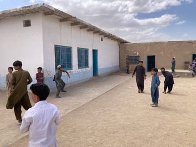 The image depicts a group of young boys playing in the courtyard of a school in Saranan, Pakistan. The school building is a simple, single-story structure with a white exterior and blue trim. The boys are wearing traditional clothing, including shalwar kameez and turbans. They are playing a game of cricket, with one boy holding a bat and another holding a ball. The court is made of dirt and is surrounded by a low wall. The weather is sunny and clear.  The school is located at coordinates 30.532 N, 66.909 E.