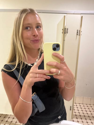 A female individual with long blonde hair is depicted from the waist up in a mirror selfie, taken within what appears to be a public restroom in Thirroul, Australia. She wears a black t-shirt, a white multi-bead necklace, a silver bangle on her left wrist, a silver chain bracelet on her right wrist, and five silver rings distributed across both hands. A blue shoulder bag with a gold-toned buckle is visible on her left side. She holds a yellow-cased smartphone in her right hand, capturing her reflection. The background shows light-colored walls and two cream-colored restroom stall doors with silver-toned hinges and latches. The floor features a geometric pattern of light and dark square tiles. A portion of a white ceramic sink is visible at the bottom of the frame. Her facial expression shows a slight smirk, with her gaze directed towards the upper right of the reflected image.
