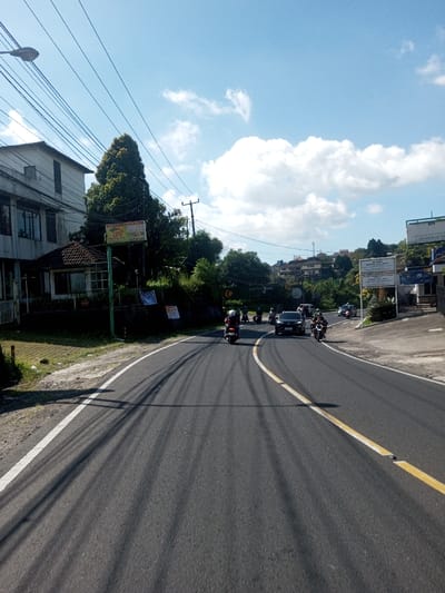 The image displays a curved asphalt road in Ban Phonhxavanh, Laos, under a bright, partly cloudy blue sky. Long, parallel shadows, likely from overhead utility wires, stretch across the road from left to right, indicating a high sun angle.

On the left side of the road, a two-story building with a light-colored facade and dark-framed windows is visible, adjacent to a smaller, single-story structure with a dark-tiled roof. A large green tree stands behind these buildings. A network of black utility wires is strung across the upper left of the frame, supported by poles, including one on the far left with a street light fixture. A yellow-green rectangular sign, partially obscured but displaying text consistent with Indonesian language (e.g., "LSG," "Jl. Raya Puputan 152," a phone number), is mounted on a pole near the buildings. Below the sign, an embankment covered with green foliage features several hanging banners or tarpaulins.

The road itself is marked with a solid white line along its left edge and a double yellow line (one solid, one dashed) on the right side of the visible lanes. Traffic consists primarily of motorcycles and at least one car, all moving away from the viewer. In the immediate foreground, a dark-colored scooter with a rider wearing a dark helmet and clothing is positioned on the road. Behind it, a black car and several other motorcycles with riders are visible further along the curve. On the right side of the road, more buildings are situated on a sloping hillside. Multiple white signs with dark text, also consistent with Indonesian language (e.g., "MASAR PENGELUARAN OBAT," "Dr. YANITA, S.H.," "Dr. JENDRA, S.H."), are prominently displayed.