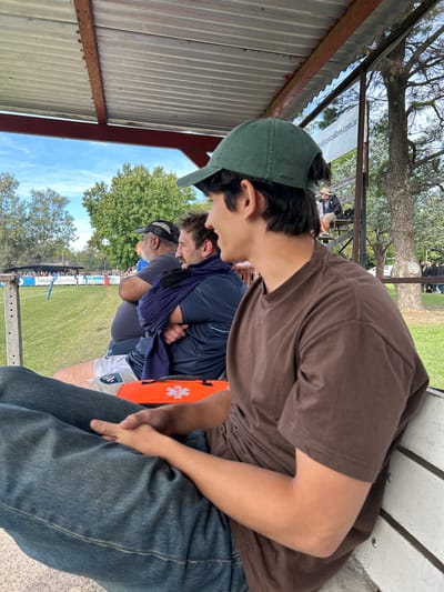 The image shows three men sitting on a bench under a metal roofed structure. The foreground figure wears a brown t-shirt and blue jeans and a dark green baseball cap. The middle figure wears a dark blue shirt and has a dark blue scarf around his neck and a small orange bag is next to him. The background figure wears a grey t-shirt and a black baseball cap. A grassy field and trees are visible behind them and some spectators. A white banner with black writing is also visible. The location is in Moreno, Argentina.