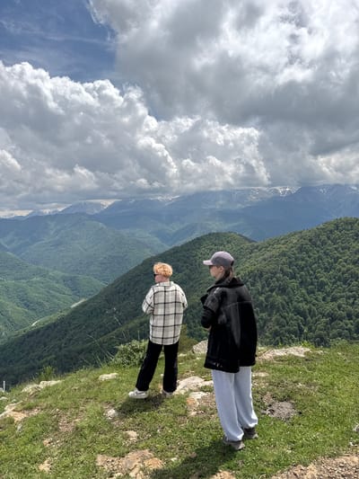 Two individuals stand on a grassy precipice overlooking a vast, verdant mountain range. The person on the left, with short, reddish hair, wears a black and white checkered long-sleeved shirt, black pants, and white sneakers. The individual on the right, facing away from the viewer, wears a grey baseball cap, a black jacket, and light grey sweatpants with grey athletic shoes. Their backs are to the camera, suggesting they are observing the expansive landscape.

The environment is a mountain vista with layers of densely forested slopes stretching into the distance. Snow-capped peaks are visible on the far horizon, under a dramatic sky filled with large, white, cumulus clouds interspersed with patches of blue sky. The terrain in the foreground is uneven and covered with green grass and scattered rocks, indicating a natural, undeveloped area. The scene is likely located in or near the city of Nizhnyaya Teberda, Russia, known for its mountainous geography and natural beauty. The lighting suggests it is daytime, with the sun illuminating the scene from above. There are no apparent interactions between the two individuals, each appearing engrossed in their view of the landscape.