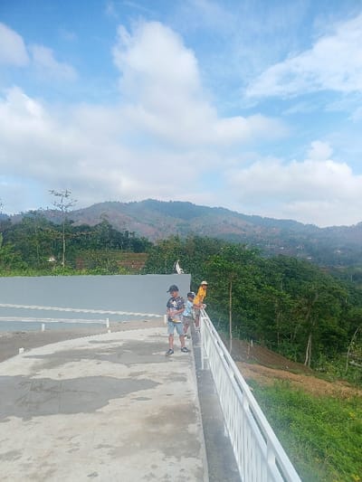 The image presents a daytime scene from an elevated concrete platform overlooking a vast, mountainous landscape, likely in Pangalengan, Indonesia. In the foreground, a smooth, grey concrete surface forms the platform, with a white metal railing running along its right edge.
Three individuals are standing near this railing, observing the view. On the left, a young person wearing a black and white patterned t-shirt and shorts stands casually. Beside them, a smaller child in a light blue top is leaning slightly over the railing. Further to the right, an adult or older youth in a yellow shirt and a light-colored hat is also leaning on the railing, looking out at the scenery.
Behind the platform and the people, a grey wall or building structure is partially visible. The midground is characterized by a steep, lush green hillside covered with dense trees and vegetation, extending downwards from the platform.
The background features a series of rolling hills and mountains that stretch across the horizon. These mountains are covered in a mixture of dark green foliage and lighter patches, possibly indicating terraced farming or areas with less dense vegetation. Hints of small structures are visible on the distant hillsides.
The sky is bright blue with scattered, fluffy white clouds, indicating clear and sunny weather. The overall impression is one of a tranquil, scenic overlook, with the individuals enjoying the panoramic natural beauty of the mountainous region. No visible text is present in the image.