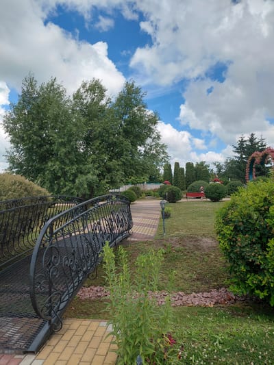 The image depicts an outdoor scene in the city of Kovalivka, Ukraine, likely a park or garden area. In the foreground, a decorative black metal bridge with intricate scrollwork spans across a paved walkway made of yellow and red rectangular bricks. To the left of the bridge, lush green foliage is visible. Beyond the bridge, a neatly manicured lawn is dotted with various shrubs and trees. In the middle ground, a person is seated on a red bench situated on the grass. Further in the background, more trees and a decorative floral archway can be seen against a bright blue sky filled with fluffy white clouds. The overall atmosphere suggests a pleasant, well-maintained outdoor recreational space.