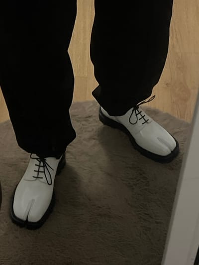 The image showcases a person wearing black pants and white, split-toe shoes. The shoes have black laces and a thick, black sole. One foot is positioned on a wooden floor, while the other rests on a gray, textured rug. The photo is taken from a slightly high angle, providing a clear view of the footwear and lower portion of the outfit. The lighting is subdued, creating a soft contrast between the colors and textures in the image.