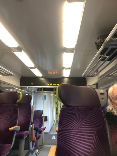 This image displays the interior of a French regional train (TER) carriage, consistent with travel in the vicinity of Dettwiller, France. The bright, overhead-lit compartment features a white, curved ceiling equipped with multiple elongated fluorescent light fixtures. On the right side, a metal, slatted overhead luggage rack contains a dark, folded item, possibly a jacket or bag. The seating comprises rows of purple upholstered chairs with a radial geometric pattern and dark purple headrests; some seats exhibit yellow frame accents or grab handles. An illuminated digital display panel, centrally located above the aisle, shows the partial French text "Ce TER Fluo est". On the far right, the upper portion of an elderly individual's head and shoulder is visible, showing light-colored hair and a dark garment with a fur-like collar. In the mid-ground on the left, a passenger's feet, shod in red and dark blue footwear, are resting on the back of an adjacent, unoccupied seat. Yellow vertical support poles or grab bars are positioned along the central aisle, which recedes towards a doorway leading to another section of the train.