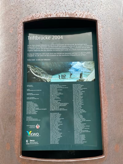 The image presents a rectangular information sign, set within a heavily rusted and pitted metal frame, indicative of weathered corten steel. The sign itself features a dark green background with white text and a color photograph.

The prominent title at the top reads "Triftbrücke 2004." The accompanying German text describes the original Trift Bridge, built in 2004, as a 102-meter-long, 1720-meter-altitude, Nepalese-style suspension bridge, a pioneering structure in the Alpine region. It explains that the bridge was constructed to access the Trifthütte after glacier retreat and was replaced by a new bridge in 2009. The text also states that the 2004 bridge's construction was made possible by numerous sponsors and friends to establish a successful tourism history, concluding with "Vielen Dank – es lebe die Triftbrücke!" (Many thanks – long live the Trift Bridge!).

Below the main text, a horizontal photograph depicts the Trift Bridge spanning a turquoise glacial lake. A large glacier is visible in the background, set against rugged mountainous terrain. Several individuals are shown walking across the bridge from left to right.

The lower part of the sign lists contributors under various headings: "Bauherrschaft" (Client: SAC Sektion Bern), "Projekt" (Project: Hüttenkommission der SAC Sektion Bern), "Projektleitung" (Project Management: Walter Brog, Hüttenchef Trifthütte), "Statik, Konstruktion" (Statics, Construction: Ingenieurbüro Hans Pfaffen, Chur), and "Montageteam" (Assembly Team), which includes names and roles such as "Schlosser" (locksmith), "Bergführer" (mountain guide), and "Servicemonteur" (service technician).

Further down, the "Trägerschaft" (Sponsorship/Support) section displays logos for "Schweizer Alpenclub SAC," "KWO Grimselstrom," and "beco Berner Wirtschaft Economie bernoise." A comprehensive list titled "Sponsoren und Freunde" (Sponsors and Friends) fills the right side of the sign, enumerating numerous individuals and organizations, with many entries specifying locations such as Meiringen and Innertkirchen.

The sign is an outdoor informational display detailing the history and support behind the Trift Bridge project, situated in what is identified as the Innertkirchen area of Switzerland.