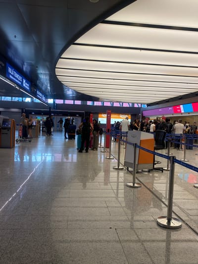 The image depicts a terminal in Ezeiza, Argentina, identifiable by signage in Spanish. A crowd of people is lined up behind stanchions, likely waiting at check-in counters. Many have luggage with them. The terminal has a curved wall design with blue and white signage in the lower portion. The floor is tiled with a neutral-toned, speckled design. Above, there is a white, paneled ceiling with linear recessed lighting.