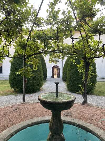 The image depicts a courtyard in Balerna, Switzerland. In the foreground, a weathered stone fountain with turquoise water is partially visible. Water streams from a central spout and spills over the fountain's rim into the pool below. The fountain is surrounded by a gravel pathway with reddish-brown pebbles.

Behind the fountain, two conical cypress trees stand symmetrically, flanking a gravel path that leads towards an arched doorway in the background. Overhanging the scene is a trellis covered in lush green grapevines, their leaves creating a dappled canopy. The walls of a building, painted white, are visible behind the cypress trees, with a barred window on the left and the arched doorway on the right. The overall impression is one of a serene, manicured garden space. There are no people visible in the image.