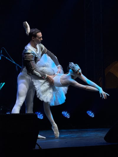A male ballet dancer in a decorative jacket and white tights supports a female dancer in a white tutu and tiara. She has one arm raised gracefully, and one leg extended, the other is bent at the knee behind her. The dancers are illuminated by blue stage lights. A microphone stand is near their feet, and a speaker is in the foreground. The background is dark.