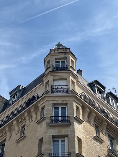 The image shows the corner of a multi-story building with a light-colored stone facade and a dark mansard roof. The building has several windows, some with white shutters and black wrought-iron balconies. At the very top, there is a small, domed structure with a finial. The sky above is a clear blue with a thin contrail. The building appears to be located in a European city, likely Paris or a nearby suburb, given the architectural style. There are no people visible in the image. The perspective is looking up towards the corner of the building.