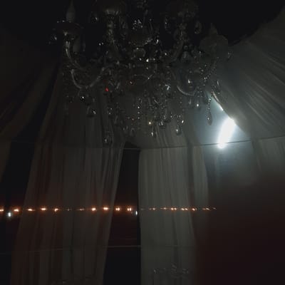 The image depicts a dimly lit indoor scene, likely a banquet hall or event space, with a grand chandelier as the central focal point. The chandelier is ornate, featuring multiple tiers of crystals that reflect the available light. Surrounding the chandelier are draped white fabrics, possibly curtains or decorative coverings, which create a soft, ethereal ambiance. In the background, a series of small, warm lights are visible, strung horizontally, adding a festive touch to the setting. A bright, diffused light source is also present to the right of the chandelier, potentially a window or an artificial light, creating a lens flare effect. The overall impression is one of elegance and celebration. The image does not contain any discernible elements that would allow for the identification of its location in the city of Sosnovka, Kyrgyzstan. There are no people, specific landmarks, or contextual clues that would indicate its geographical placement within that city.
