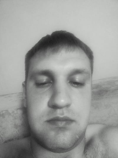 The image is a close-up, black and white, frontal selfie of a man's face. He has short, dark hair and his eyes are closed. His face is looking down, and the lighting suggests an indoor setting. A thin chain necklace is visible around his neck. The background is out of focus and appears to be a textured surface, possibly a wall or furniture. There is no identifiable information in the image that would place it in Sorochinsk, Russia.