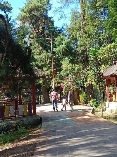 A sunny, clear day in Gundaling 2, Indonesia. Three people walk on a paved pathway through a park. The person furthest back is a woman wearing a headscarf and a grey outfit. Next to her is a young boy wearing a white shirt and shorts. In front of them is a man in a red and white checkered shirt and blue jeans. The pathway is paved with square tiles and is bordered by greenery. To the left of the pathway are some structures with colorful decorative panels, and further back are large trees and a tall, thin pole. To the right, there is more lush vegetation and a small red structure. Sunlight casts shadows across the path, indicating it is likely mid-morning or afternoon.
