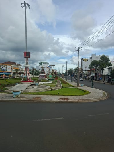 The image shows a street intersection in the city of Bao Loc, Lam Dong province, Vietnam. In the foreground, there is a wet asphalt road with white lane markings. A curved concrete sidewalk with decorative paving stones lines the edge of the road. Beyond the sidewalk is a well-maintained green median with manicured grass and small blue planters.
Dominating the left side of the median is a tall, silver lamppost with multiple lights at its apex. A red and white structure is attached to the lamppost, featuring a digital display screen. Further into the median, a decorative circular monument with a tiered, floral design is visible.
The street is lined with buildings on both sides. On the left, a building with an orange roof has a large advertisement featuring people. Further down, a multi-story building with a white facade and blue accents has a sign that reads "SANDALS HOTEL". Other businesses with various signage are present along the street, including what appears to be a post office.
Several vehicles are on the road, including cars and motorcycles, indicating moderate traffic. A few people are visible, mostly on motorcycles. The sky is overcast with fluffy white and grey clouds, suggesting it may have recently rained or is about to. The overall environment appears to be a typical urban street scene in a Vietnamese city.