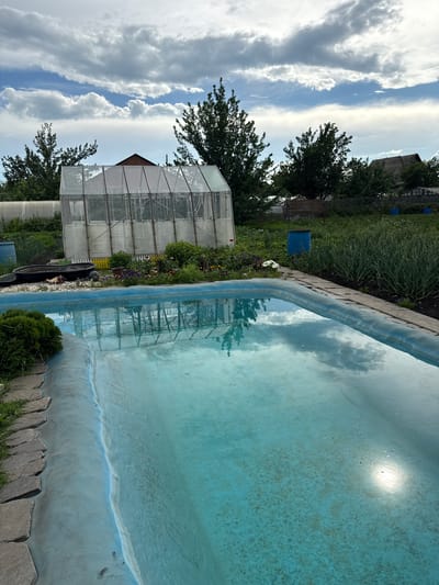 The image depicts a scene in Bogdanovich, Russia, featuring an empty swimming pool with turquoise water. The pool's edge is lined with decorative stones and grass. In the background, a greenhouse with a wooden frame and transparent covering stands amongst lush green foliage, including trees and what appears to be a vegetable garden. Several blue barrels are visible near the greenhouse and further into the background. The sky above is a dynamic mix of white, fluffy clouds and patches of blue, suggesting partly cloudy weather. Sunlight reflects off the water's surface, creating bright spots and a sense of warmth, indicating it is likely daytime. The overall impression is one of a rural or semi-rural setting, possibly a backyard or a communal garden area, with elements of both recreation and cultivation. No people are visible in the frame. The image is oriented vertically, suggesting it was taken with a phone camera held in portrait mode.