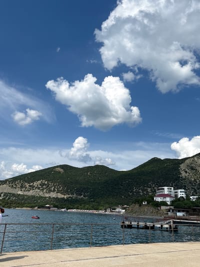 The image displays a coastal scene in Bol'shoi Utrish, Russia, under a blue sky with scattered cumulus clouds. In the foreground, a light-colored concrete pier or walkway is visible, bordered by a multi-bar metal railing that extends horizontally across the lower frame. The midground features a body of water with discernible ripples. A small red inflatable boat carrying two individuals is positioned in the water towards the left. Further right, a concrete pier structure with railings projects into the water from the coastline. The shoreline along the midground includes a populated beach area with numerous sun loungers and umbrellas. Behind the beach, several buildings are visible, including white multi-story structures with red roofs on a rocky outcrop to the right. The background is dominated by two large, verdant mountains covered with dense tree foliage, sloping down to the water's edge. Patches of exposed rock are visible on the mountain slopes. The sky above is clear blue, containing various white clouds ranging from small, wispy cirrus-like formations to large, fluffy cumulus clouds, with a significant cloud mass occupying the upper right quadrant.