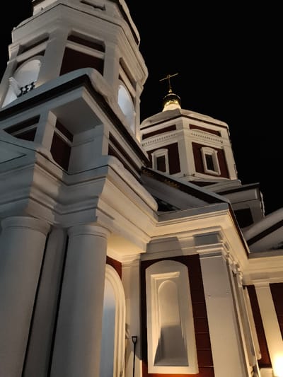 A nighttime photograph captures an illuminated religious edifice in the city of Im. Gastello, Russia. The structure is predominantly white with dark red (maroon) accents, featuring multiple tiers, classical columns, pilasters, and cornices. On the left, a section of a bell tower with arched openings is visible, exhibiting dark interiors. To the right and slightly recessed, a smaller, square-based tower culminates in a golden dome surmounted by an Orthodox cross. The building is intensely lit by artificial light sources, creating stark contrast against the dark, featureless night sky. No human subjects are present. A dark, long-handled implement, likely a shovel, rests vertically against the building's base on the lower right, adjacent to a shallow arched niche.