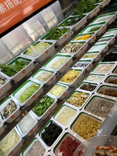 The image shows an overhead view of a food display at a restaurant in Bottrop, Germany. The display features numerous white rectangular containers filled with various ingredients, primarily vegetables and some protein options. The containers are arranged in tiered rows on a metallic surface, suggesting a self-serve buffet or a preparation area for dishes like hot pot or fondue.
Specific ingredients visible include:
- Leafy green vegetables (e.g., bok choy, spinach, lettuce)
- Cabbage and napa cabbage
- Mushrooms (e.g., enoki, shiitake)
- Broccoli florets
- Bamboo shoots
- Sliced lotus root
- Sliced potatoes and potato slices with a crinkle-cut pattern
- Tofu cubes
- Bean sprouts
- Sliced onions
- Noodles (various types, including flat yellow noodles and thin white noodles)
- Seaweed
- Raw thinly sliced beef
- Shrimp
- Fried tofu cubes
- Some cooked meat items are also visible in containers further down.
Several containers have green trim and small circular red stickers with Chinese characters, indicating the brand or origin of the food. Some labels with German text are also partially visible, such as "Bambus" (bamboo), "Brokkoli," "Holzrasling dunkel" (dark wood ear mushroom), and "Tofu Streifen dick" (thick tofu strips). An orange sign with white Chinese characters is visible at the top of the image. The overall impression is one of a well-stocked and diverse food selection, likely for a hot pot or a similar communal dining experience. No people are clearly visible in the frame, and the focus is entirely on the food presentation. The environment appears to be indoors, with a clean and organized setup.