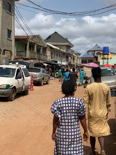 The image shows a street scene in the city of Ondo, Nigeria, located at coordinates 7.251 latitude, 5.196 longitude. There are several people walking along the street, some wearing brightly colored clothing. Some people are walking in a group. There are also several parked vehicles, including a white van, a silver car, and a brown car. There are buildings on both sides of the street, some with balconies and windows. There are many electrical wires running above the street. The street is unpaved, with dirt and gravel. There are signs of businesses and shops on the street. There is a blue water tank on a metal stand on the right side of the street. It is a sunny day. The sky is blue and partially cloudy.