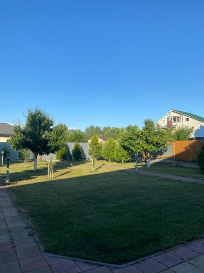 This is a vertical, eye-level, outdoor shot of a residential backyard in Volzhsk, Russia. The scene is bathed in the bright, clear light of a sunny day, with a vast expanse of brilliant blue sky dominating the upper portion of the frame. The foreground is paved with a patterned walkway made of reddish-brown and gray rectangular tiles, bordered by a low concrete edge and a well-maintained green lawn.
Several young trees and bushes are scattered across the lawn. A larger, mature tree with a white-painted trunk stands prominently in the left-middle ground, casting a shadow across the grass. Various smaller trees, some appearing to be evergreens, are also present. Pathway lights, with dark poles and lanterns, are situated at intervals along the paved walkway.
A metal fence runs horizontally across the middle ground, separating the yard from adjacent properties. Beyond this fence, a wooden fence section with vertical planks is visible on the right. In the background, residential buildings with pitched roofs are partially visible through a line of green trees. The overall impression is one of a well-kept, suburban backyard. There are no people or animals visible in the image, and no actions or interactions are depicted.